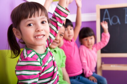 A group of diverse elementary students in a classroom, with one young girl in a striped shirt smiling and raising her hand to participate in a fun reading activity.