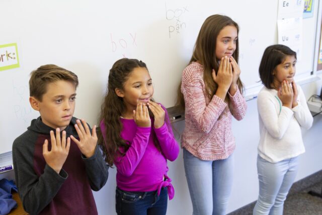 Four elementary students standing in a classroom participating in a multisensory dictation activity, using hand motions and vocalizing sounds to reinforce letter-sound relationships.