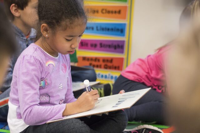A young kindergarten student smiling while holding up a small whiteboard to show the teacher their answer during a "hold-up" assessment.