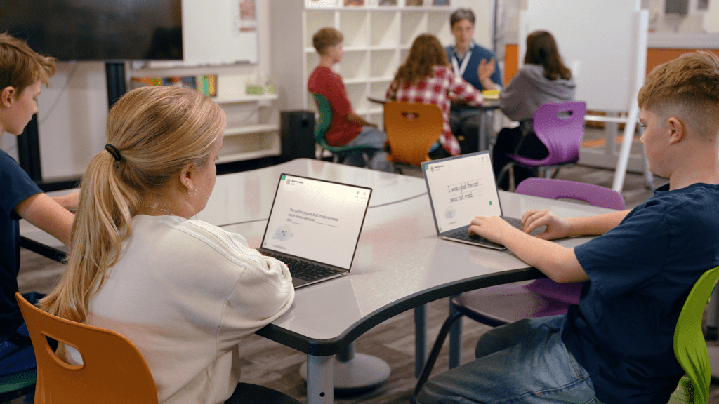 Students work independently on laptops while a teacher leads a small-group lesson.