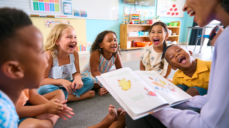 A teacher reads to her class of young students who all show excited faces.